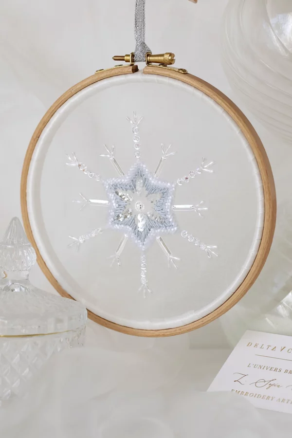 Un délicat flocon de neige brodé à la main, Bianca, présenté dans un décor d'inspiration hivernale, capturant la beauté cristalline de la nature.