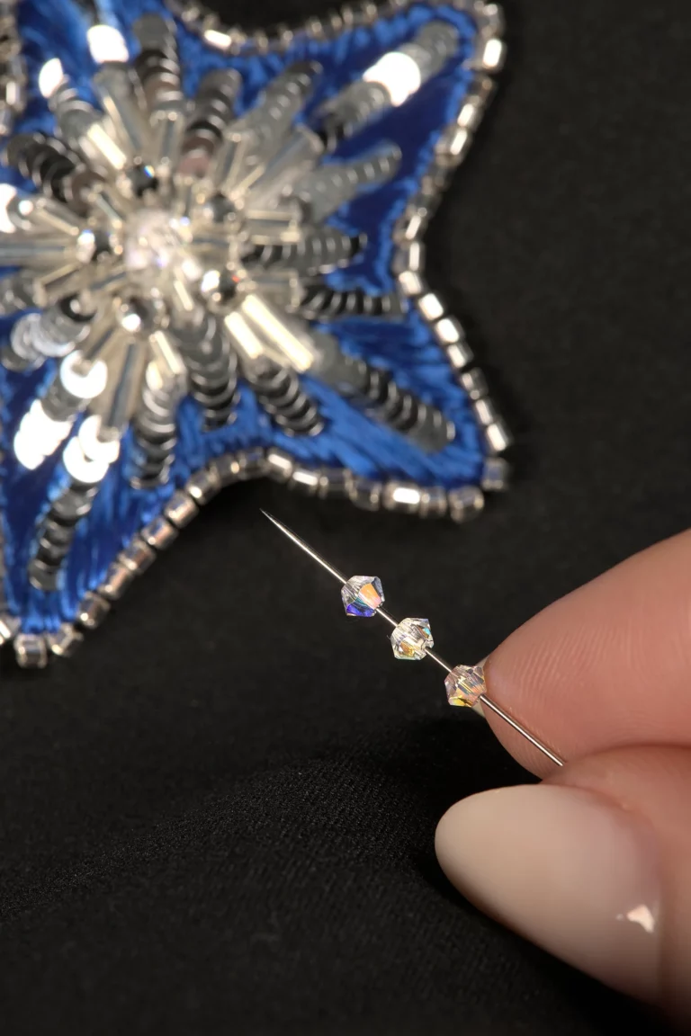 Primer plano de unos dedos sosteniendo una aguja con cristales de Swarovski, con un broche de estrella bordado de fondo, mostrando un curso de bordado de Alta Costura en línea.