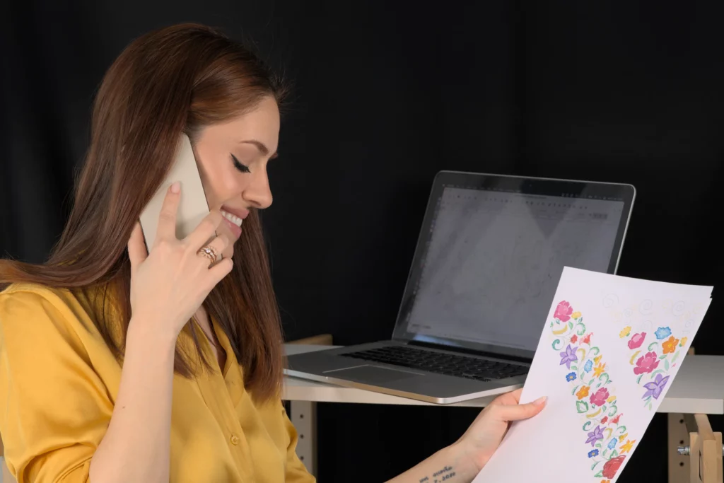 Laura S. Urbina discute d'un motif de broderie florale avec un client au téléphone, tenant le projet final avec son écran d'ordinateur en arrière-plan.