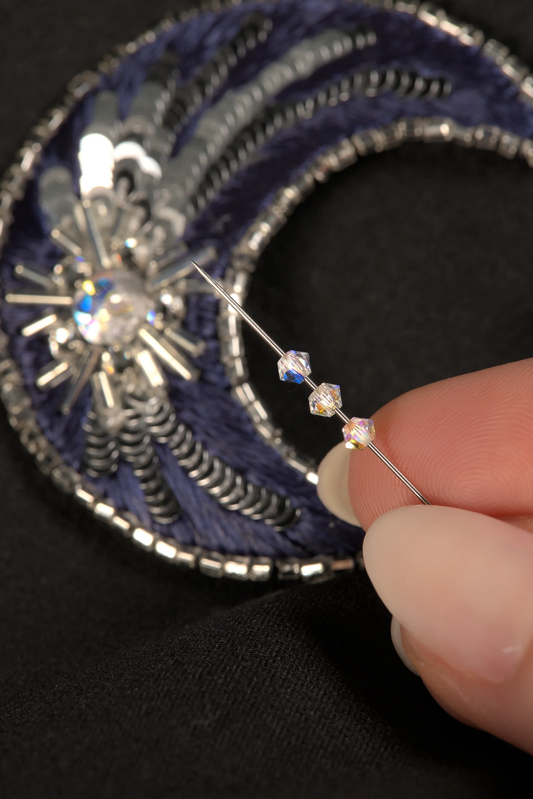 Close-up of fingers holding a needle with Swarovski crystals, with an embroidered moon brooch in the background, showcasing an online Haute Couture embroidery course.