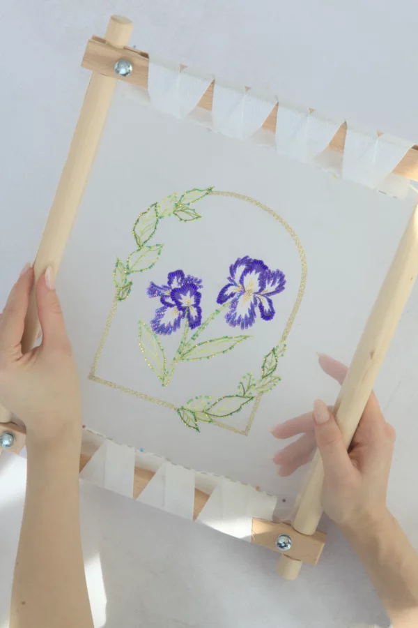 Hands holding Nereid embroidery sample with iris flowers and leaves, created using Lunéville hook technique.