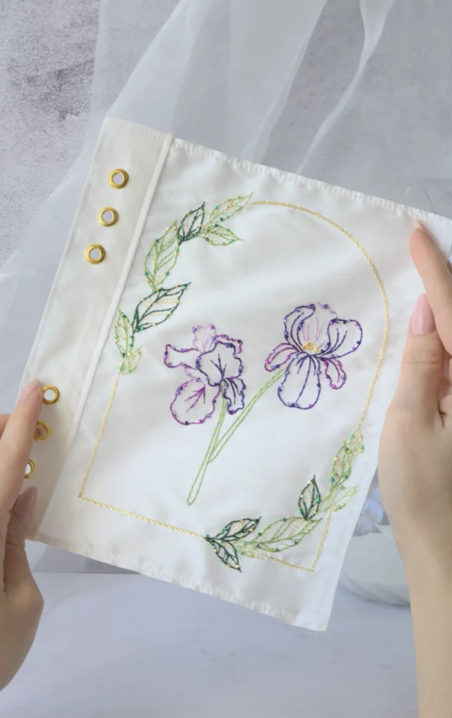 Échantillon de broderie Nereid tenu entre les mains, avec des fleurs et des feuilles d'iris, créé avec la technique au crochet de Lunéville.