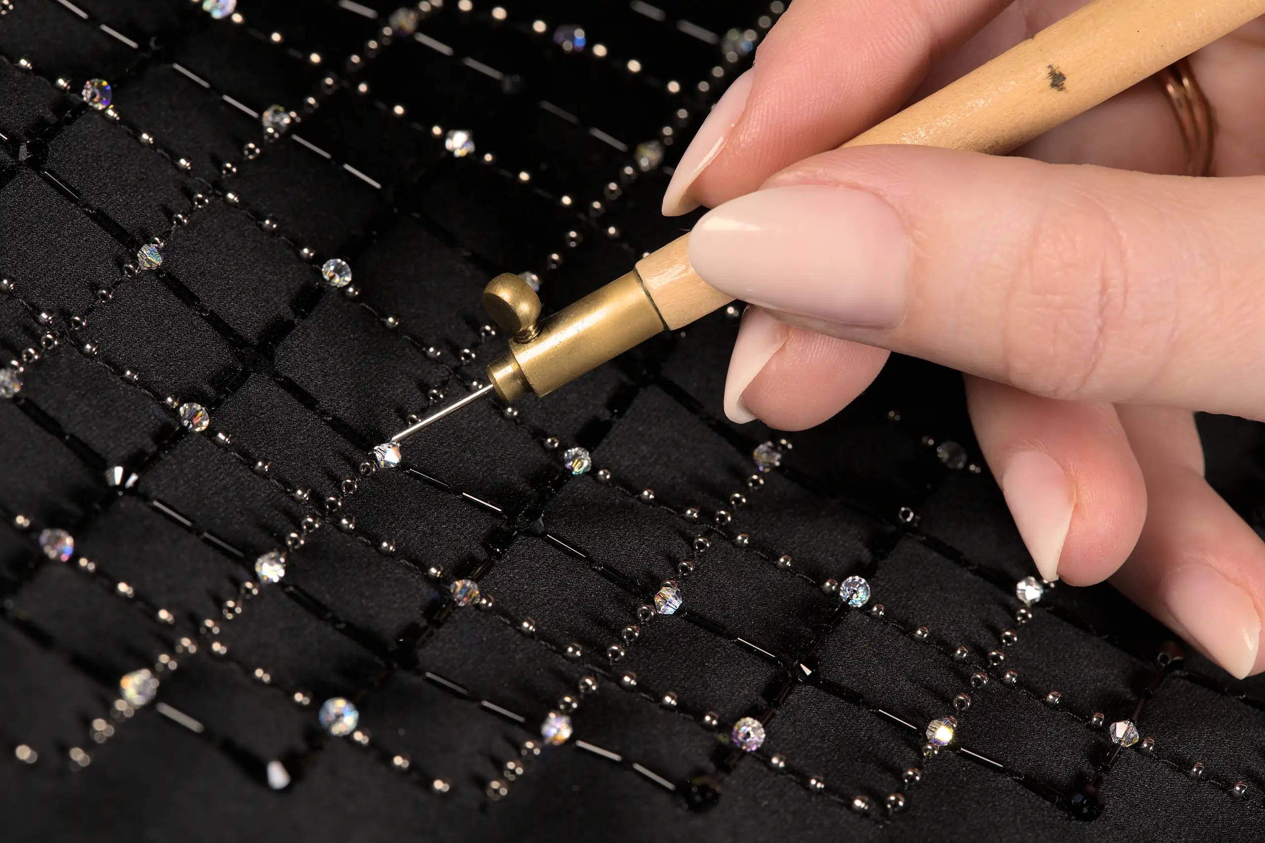 Gros plan d’un crochet de Lunéville tenu par les doigts, avec en arrière-plan une élégante grille brodée en noir, présentant l'Académie de broderie de Delta V Créations.