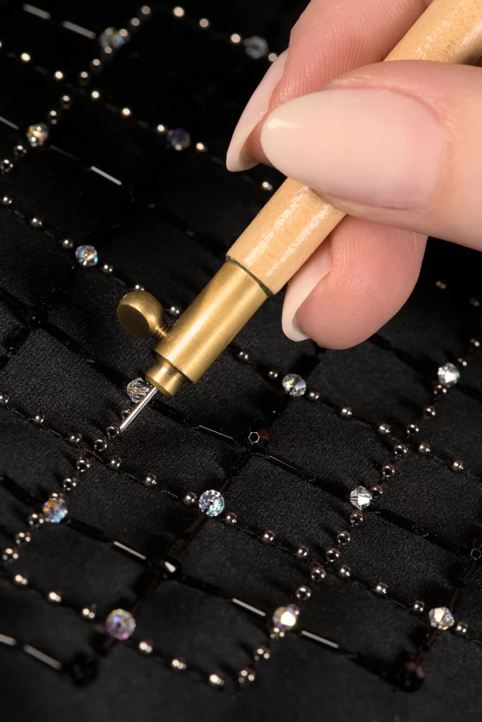Lunéville hook held by fingers, with an elegant black embroidered grid in the background, introducing the Delta V Créations Embroidery Academy.