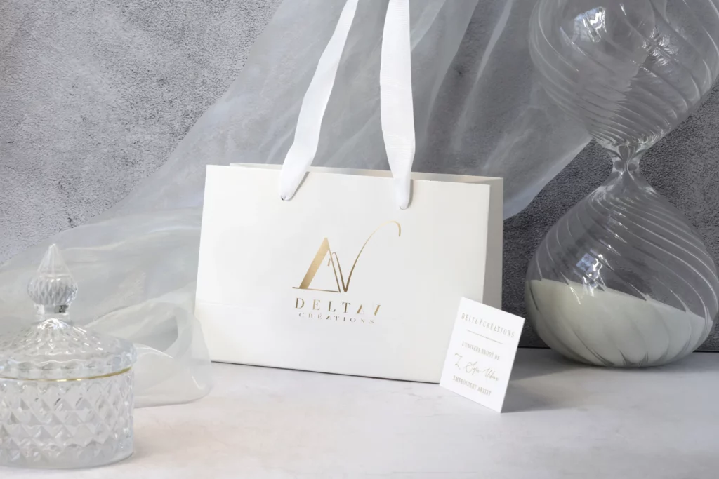 Elegant Delta V Créations luxury shopping bag with gold logo, accompanied by a refined business card and glass décor, evoking sophistication and haute couture craftsmanship.
