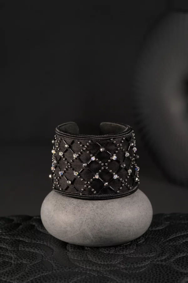 Un brazalete negro bordado con cristales se presenta sobre un expositor de cemento, resaltando el saber hacer de la alta costura francesa en una creación atemporal y elegante.