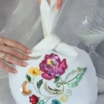 Off-white Japanese knot bag with vibrant floral embroidery, held in hand. Handcrafted in France with sequins, silk threads, and beading details.