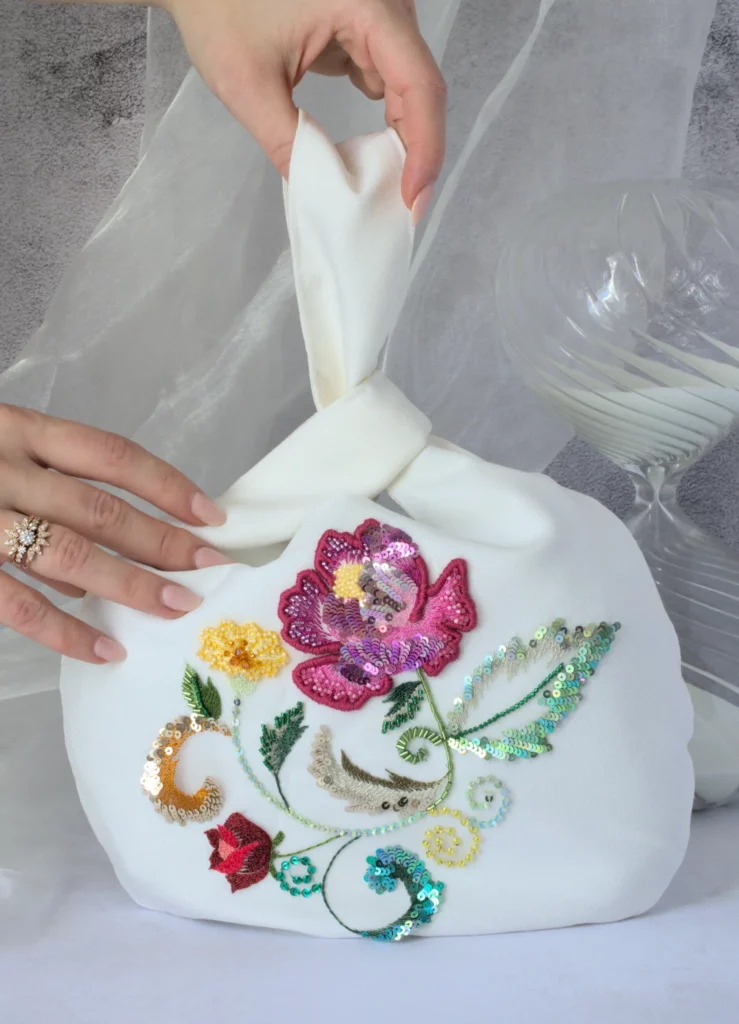 Off-white Japanese knot bag with vibrant floral embroidery, held in hand. Handcrafted in France with sequins, silk threads, and beading details.