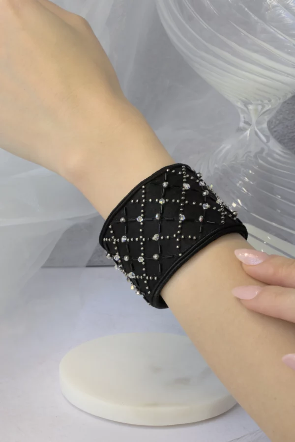 Un elegante brazalete negro bordado con cristales se presenta en la muñeca de una modelo, resaltando el bordado de alta costura y el lujo atemporal de las creaciones prêt-à-porter francesas.