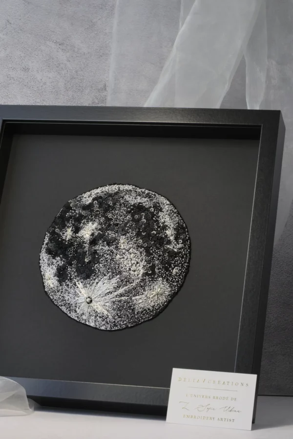 Œuvre brodée représentant la Lune, présentée encadrée avec une carte de visite de l’atelier de broderie Delta V Creations.