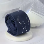 Cravate en soie roulée avec broderie des constellations d’Orion et Aquila, posée sur un socle en marbre blanc, accompagnée d’un sablier et d’un stylo en verre.