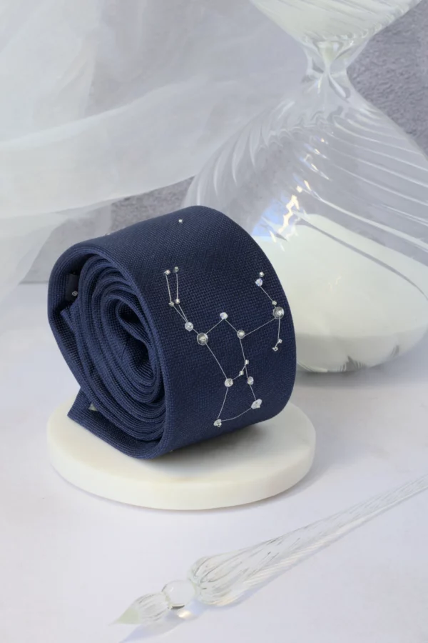 Rolled navy silk necktie embroidered with constellation stars in silver thread and crystals, styled with hourglass and pen.
