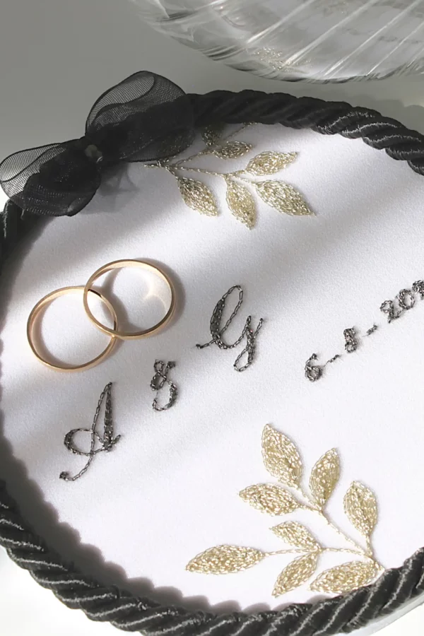 Elegant wedding ring holder with custom embroidery in black and golden tones, with two wedding bands.