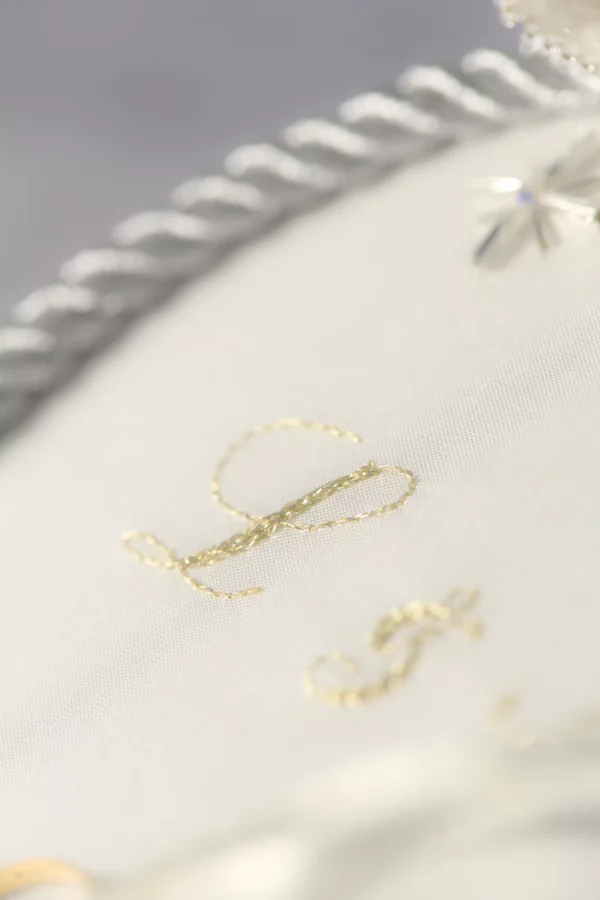 Close-up of golden embroidered initial “L” on wedding ring holder, with shimmering silver twisted cord accent in soft diagonal lighting.