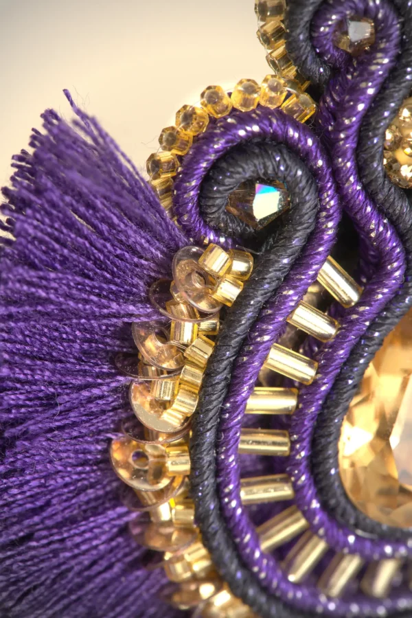 Detalle del bordado de un pendiente boho chic en violeta y oro, con trenza soutache, cristal central y pompones de algodón.