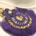Pendiente boho chic bordado a mano en violeta y oro, con cristal y pompones de algodón, sobre tela de seda color crema.