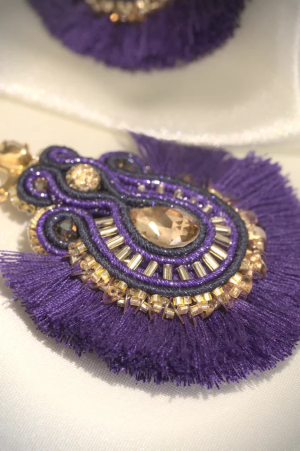 Pendiente boho chic bordado a mano en violeta y oro, con cristal y pompones de algodón, sobre tela de seda color crema.