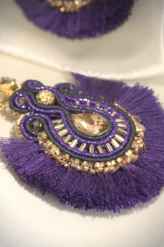 Hand-embroidered boho-chic Sunray earring in violet and gold, with crystal center and cotton fan pompoms, displayed on cream silk fabric.
