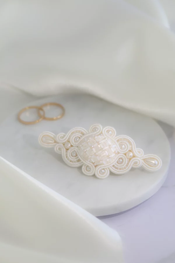 Bridal hair clip, hand-embroidered with crystals and pearls, displayed on marble beside wedding rings.