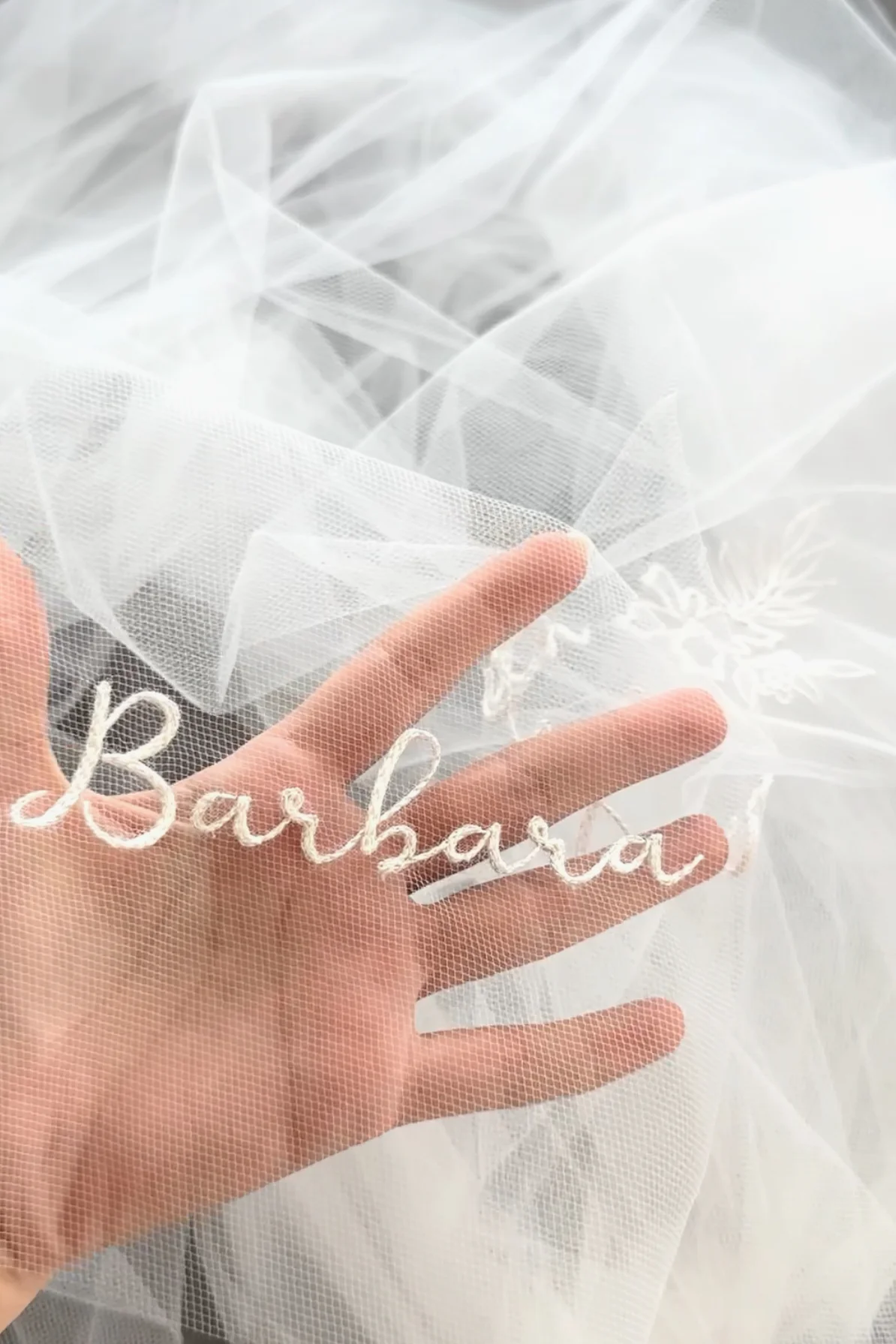 Primer plano del velo de novia bordado personalizado con el nombre de la novia, Barbara, y una mano sosteniendo la tela de tul.
