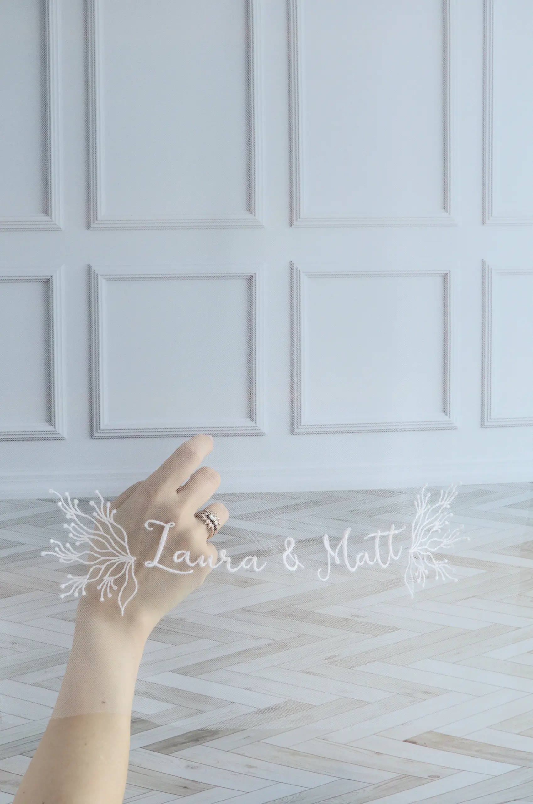 Velo de novia bordado con los nombres de los novios, mano con anillo de compromiso visible tras el tul.