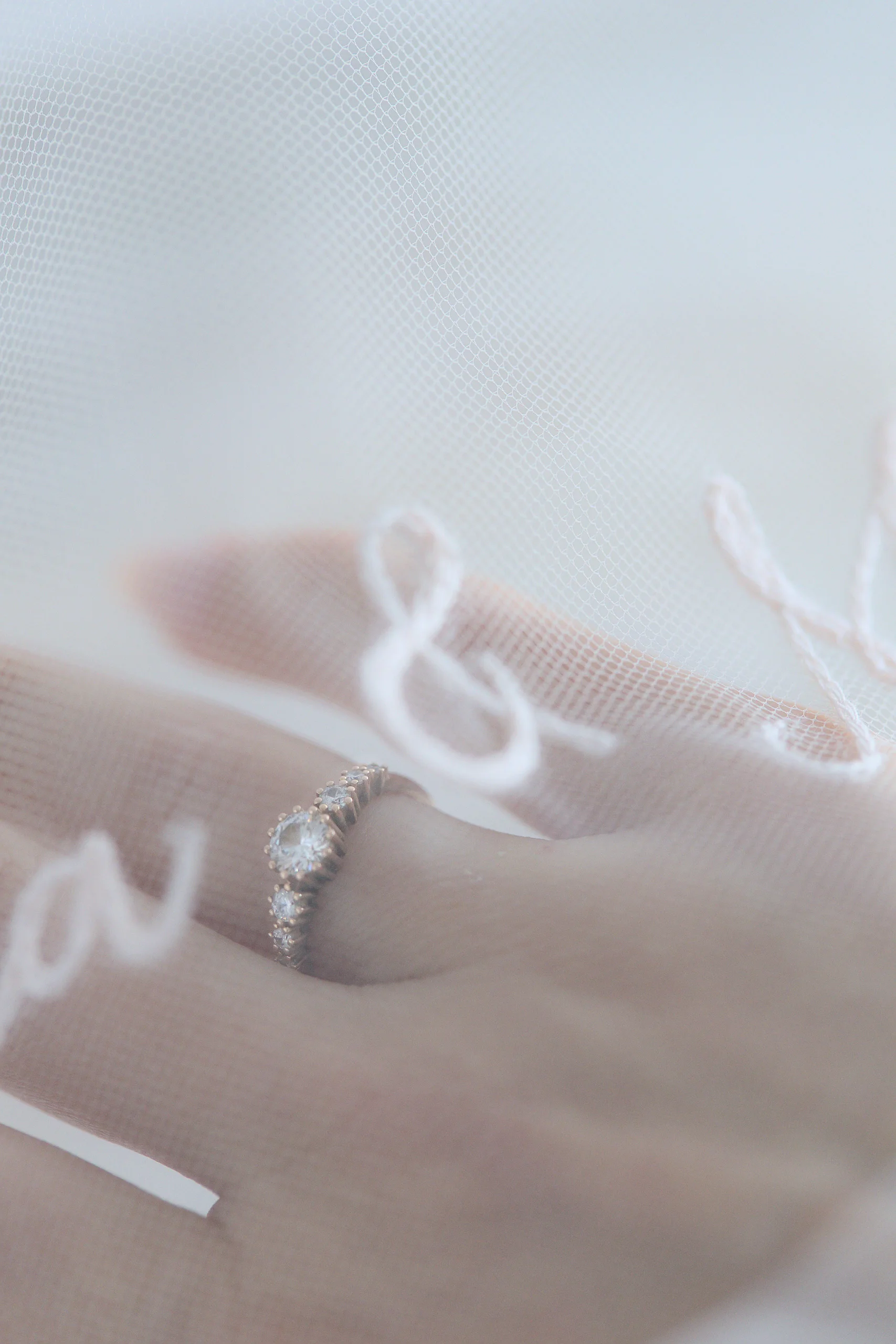 Mano con anillo de compromiso tras el velo bordado a mano, letras difuminadas visibles en el tul.