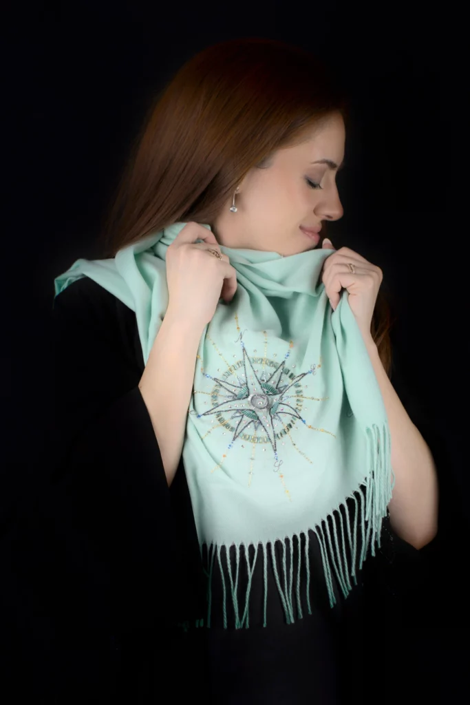 Mannequin souriant tenant l’écharpe Alcyone en cachemire turquoise avec sa rose des vents brodée à la main.
