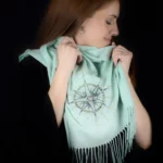 Model smiling while holding the Alcyone turquoise cashmere scarf with its hand-embroidered compass rose.