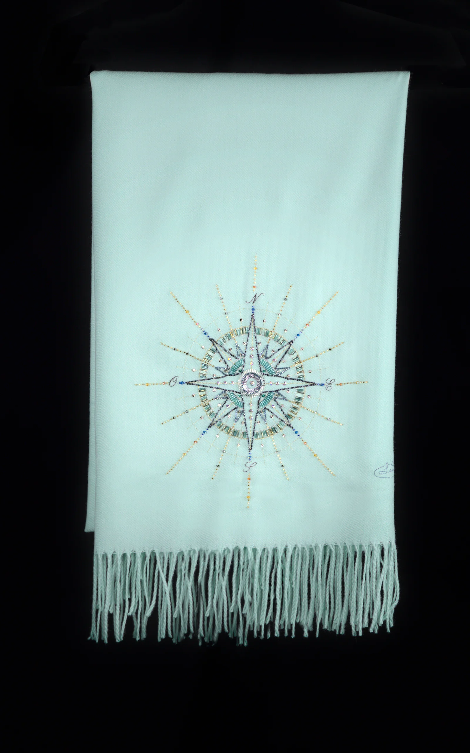 Flat lay of the Alcyone turquoise cashmere scarf showing its hand-embroidered compass rose.