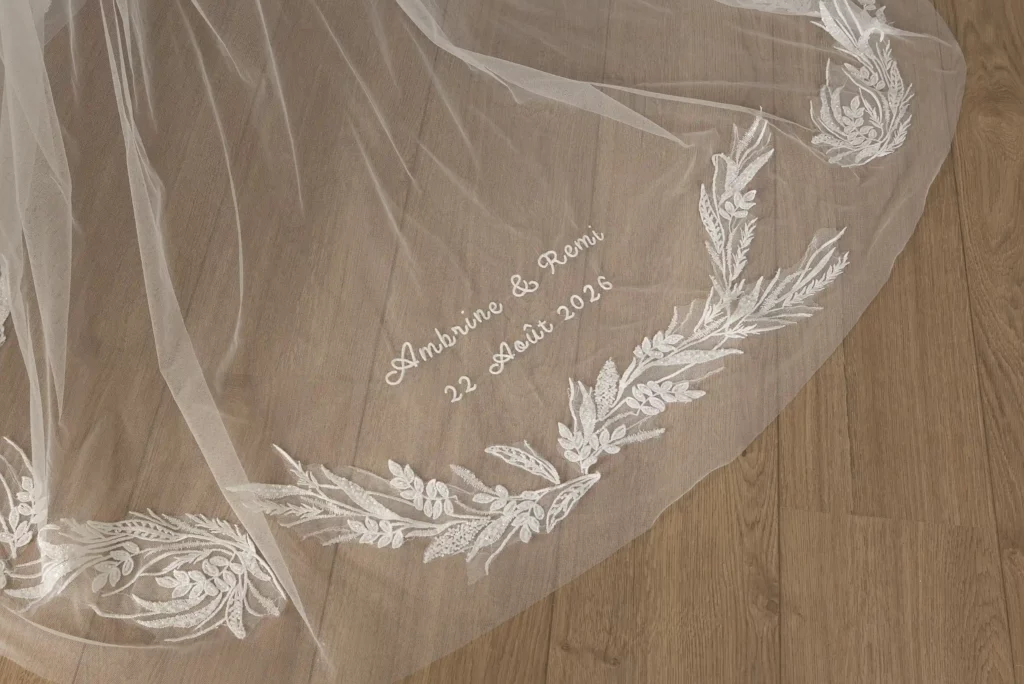 Luxury bridal veil with personalised hand embroidery of the couple’s names and wedding date, displayed over a wooden floor.