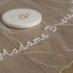 Voile de mariée brodé et minimaliste avec un prénom personnalisé, présenté avec deux alliances sur un disque en marbre.