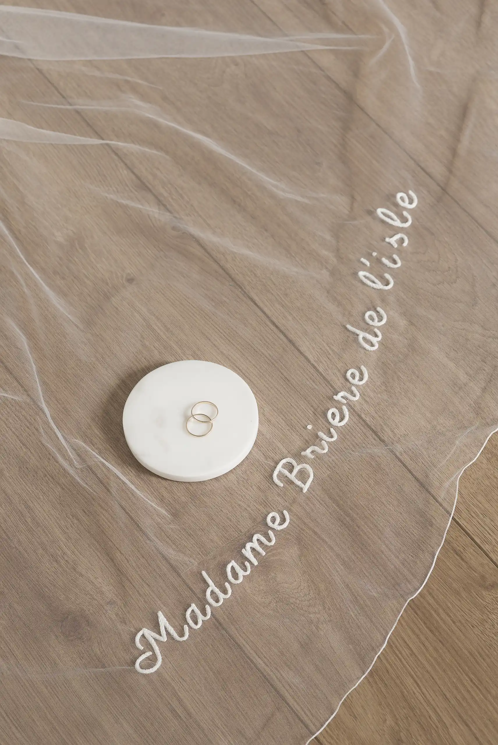 Voile de mariée minimaliste avec un nom personnalisé brodé à la main, présenté avec deux alliances sur un disque en marbre.