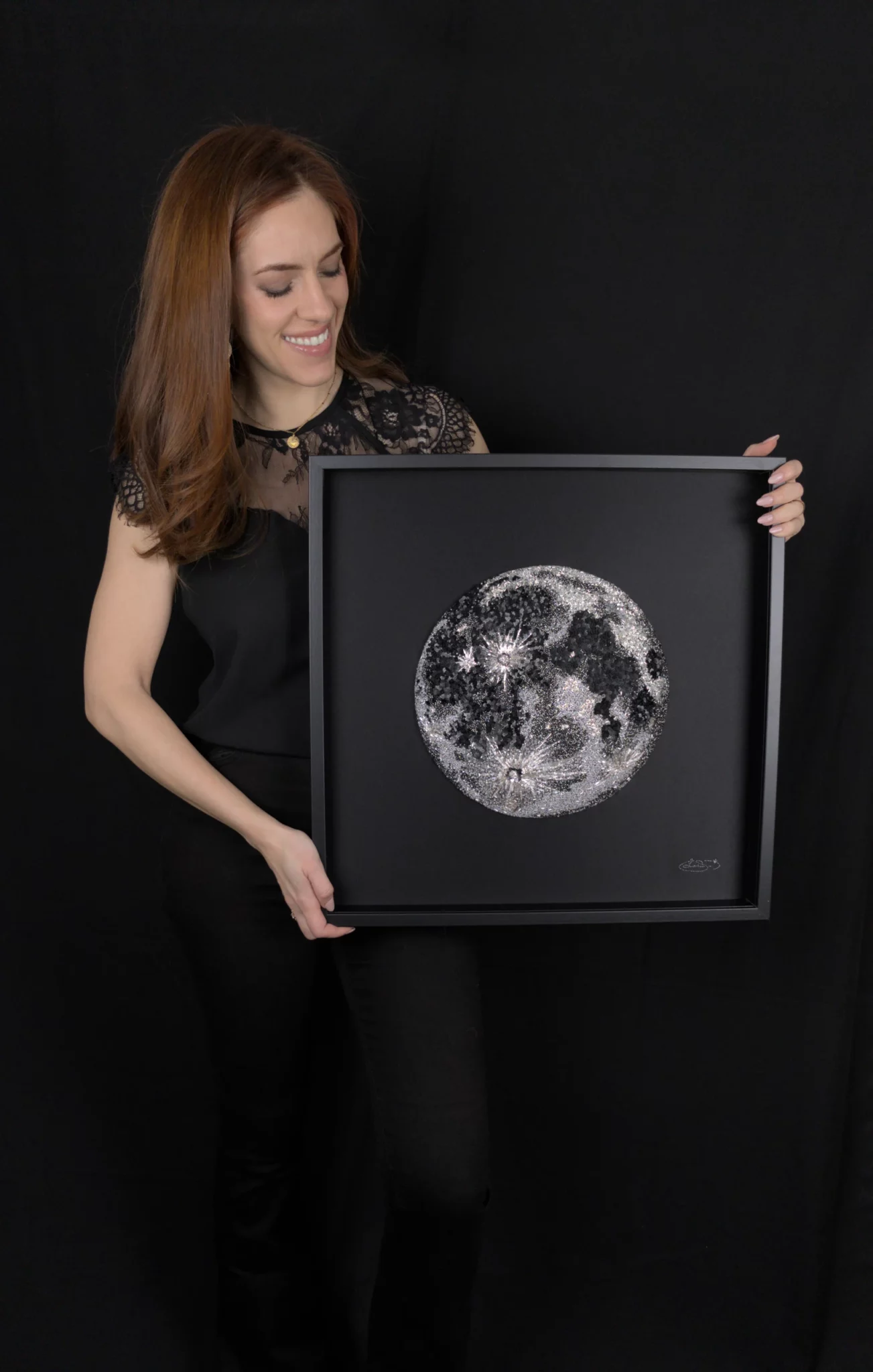 Laura S. Urbina sostiene su obra original enmarcada de una gran Luna bordada en un estudio de tonos oscuros.