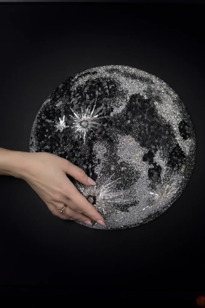 Laura S. Urbina presenting her original framed Moon artwork with her hand gently resting on its embroidered surface.